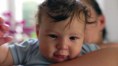 Annenin kolunda dinlenen bebek, meraklı bakışlar, yakın plan portre, parlak gözler, nazik ifade, samimi bir bağ anı, sıcaklık ve güven, barışçıl ve sevgi dolu bir bağ.