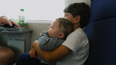 Bebek kardeşini tren koltuğunda tutan büyük kardeş, ikisi de pencereden dışarı bakıyor, barışçıl seyahat, çocukluk merakı, aile yolculuğu, şefkatli kucaklaşma, sessiz bir bağ..