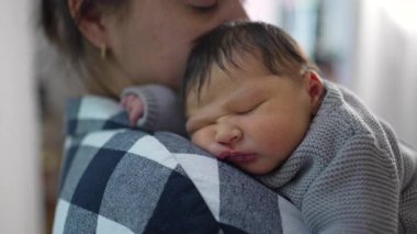 Anne, yeni doğmuş bebeğini omzunda tutarken nazikçe alnından öpüyor. Bebek rahat bir şekilde uyur, örülmüş bir süveter giyer, hassas bağlarını gösterir..