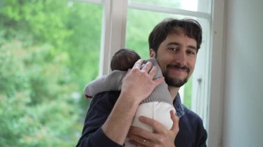 Mutlu Baba yeni doğmuş bebeğini omzunda tutuyor, kameraya sıcak bir şekilde gülümsüyor. Babacan bir sevgi ve özen gösteriyorum..