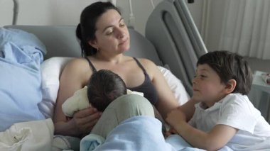 Anne kucağında yeni doğmuş bir bebeği tutarken büyük kardeş merakla ve sevgiyle ona bakıyor. Hastane yatağında geçirilen aile anları, aile birliği ve neşeyi vurgulayarak, kardeşler ve annenin sevgi dolu bakımı arasındaki bağı vurguluyor..