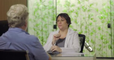 Klinik ofis ortamında kıdemli erkek hastayla yapılan istişare sırasında el hareketleriyle tedavi planını açıklayan doktor