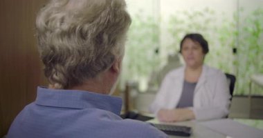 Klinikte tıbbi konsültasyon sırasında kadın doktoru dinleyen yaşlı erkek hastaya dikiz bakış açısı, bulanık bir geçmişe sahip sağlık ortamında güven ve iletişim