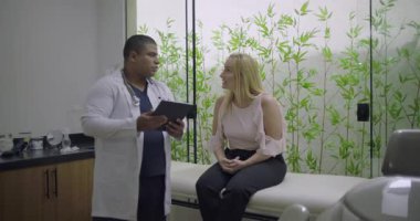 Modern klinikte randevu sırasında dijital tablet kullanan orta yaşlı bir kadın erkek doktordan tıbbi rehberlik alıyor.