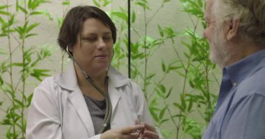 Sakin bir muayenehane ziyareti sırasında steteskopla yaşlı erkek hastaya kalp muayenesi yapan kadın doktor.