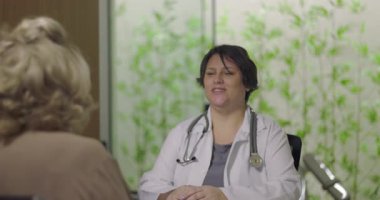 Kadın doktor, klinikteki tıbbi danışmanlık sırasında boynunda steteskop ve sakin tavırları olan yaşlı kadın hastayı dikkatle dinliyor.