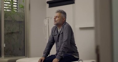 Muayene masasında tek başına oturan bir adam, danışmadan sonra duyguları incelerken sessizce yukarıya bakıyor, serumdan arkaplanda görünür bir damlama.