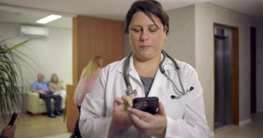 Kadın doktor, hastane lobisinde hastalar ve asansörün yanındaki resepsiyonistle gülümserken akıllı telefon kullanıyor.