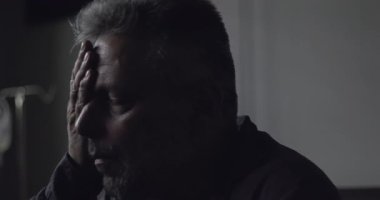 Klinikte oturan, gözleri kapalı, elleri yüzü koyun oturan, duygusal belirsizlik anında sessizce acı ve yorgunluk çeken orta yaşlı bir adamın yakın çekimi.