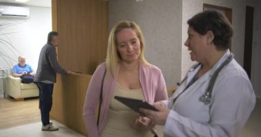 Klinik koridorunda kadın doktorla dijital sağlık bilgilerini gözden geçiren kadın, her ikisi de resepsiyon alanının yakınındaki sakin bir konuşma sırasında ekrana odaklanmış durumda.