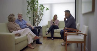 Tıbbi bekleme odasındaki hastalar rahat bir atmosferde oturuyorlar, bazıları konuşuyor ve diğerleri de telefonları kontrol ediyor, parlak klinik salonunda.