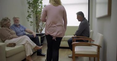 Klinikteki bekleme salonunda yürüyen bir kadın, yaşlı bir çift ve orta yaşlı bir adam da dahil olmak üzere oturmakta olan hastalara doğru yürüyordu.