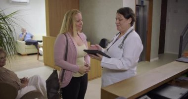 Kadın doktor, klinik resepsiyonunun yakınında kadın hastayla konuşurken, sıcak sağlık ortamındaki tablet hakkındaki bilgileri gözden geçirirken el ele verip