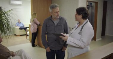 Adam, klinik koridorunda kadın doktorla konuşurken rahatladığını ifade ediyor. Elleri göğsünde gülümsüyor. Sağlık hizmetleri tartışmaları sırasında güven verici haberler alıyor.