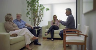 Yaşlı çift, klinik bekleme salonunda otururken sohbet ediyorlar. Etrafı diğer hastalarla çevrili. Sessiz tıbbi salondaki telefonları kullanıyorlar.
