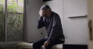 Orta yaşlı bir adam, boş bir muayenehanede gergin bir duruşla öne eğiliyor. Zor bir muayeneden sonra, yüzündeki gerginlik ve yorgunluk görülebiliyor.