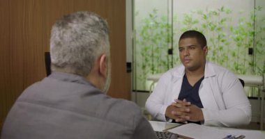 Tıbbi konsültasyon sırasında orta yaşlı bir hastayı dinleyen erkek doktor elleri masanın üstünde, arka planda bitkilerle klinikte oturuyor.