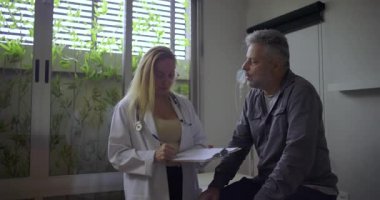 Orta yaşlı erkek hasta tıbbi danışma odasında dikkatle dinlerken kadın doktor panoya yazı yazıyor.