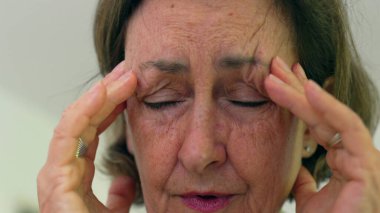 Kıdemli kadın gözlerini sıkıca kapatıyor ve iki eliyle tapınaklara masaj yapıyor. Acıyı ya da stresi azaltmaya odaklanıyor. Kırışıklıklar ve ifade, duygusal yükü vurgular