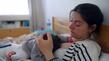 Anne, uyuyan bebeği göğsünde yatağında dinlenirken, gözleri bir an için sessiz bir yansımayla kapanıyor. Anne ve bebek arasında paylaşılan huzur ve huzur.