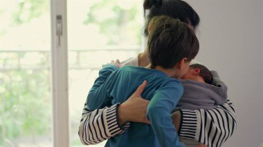 Anne hem yeni doğan hem de büyük çocuğu kucaklıyor, hassas bir aile anında kardeşler arasındaki sevgi ve ilgiyi dengeliyor, anne olmanın önemini vurguluyor.