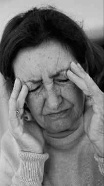 Yaşlı kadın gözlerini kapatıp şakaklarına masaj yapıyor, şiddetli baş ağrısı belirtileri gösteriyor. Onun acı ifadesine odaklan.