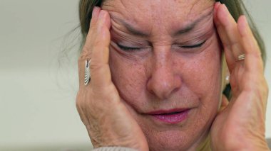 Yaşlı kadın yakın plan, gözleri kapalı, iki eli şakaklarına bastırıyor, derin kırışıklıklar ve acı dolu bir ifade.