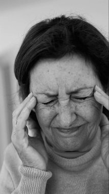 Yaşlı kadın gözlerini kapatıp şakaklarına masaj yapıyor, şiddetli baş ağrısı belirtileri gösteriyor. Onun acı ifadesine odaklan.
