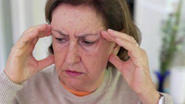 Büyük stres altındaki yaşlı kadın, parmaklarıyla tapınaklara masaj yapıyor, gözleri odaklı, yoğun endişe ve zihinsel yük altında. Detaylı ifade, duygusal gerginliğin etkisi veya fiziksel acı