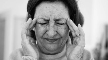 Görünür bir sıkıntı içindeki yaşlı kadın, iki eliyle yüzünü buruştururken, gözleri sımsıkı kapalı, yoğun bir şekilde ağrı kesiciye odaklanmış. Kırışıklıklar ve yüz ifadesi, baş ağrısı veya stres