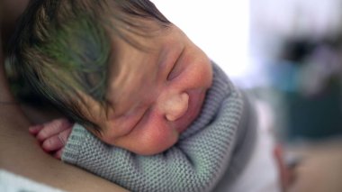 Yeni doğan bebek, somurtkan bir ifadeyle annesinin göğsünde dinleniyor. Bir anlık rahatsızlık ve annelik bakımı yakalıyor..
