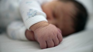 Yeni doğmuş bir bebeğin elinin yakın çekimi, uyurken usulca kıvrılmış, huzurlu bir ortamda ilk yaşamın masumiyetini ve kırılganlığını simgeliyor.