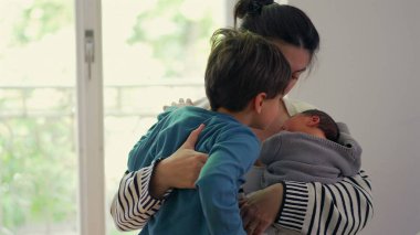 Anne hem yeni doğan hem de büyük çocuğu kucaklıyor, hassas bir aile anında kardeşler arasındaki sevgi ve ilgiyi dengeliyor, anne olmanın önemini vurguluyor.
