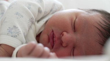 Serene uyuyan bebek, beyaz tulum içinde, bebeğin yüzünün ve elinin yakın çekimi, erken çocukluğun yumuşaklığını ve saflığını yakalıyor.