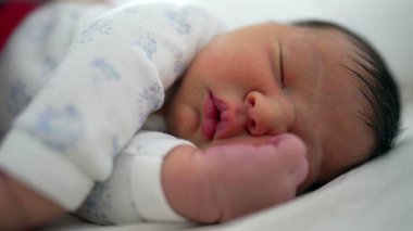 Yeni doğmuş bebek bir yatakta huzur içinde uyuyor. Nazik, rahat bir yüz ifadesi ve bir eli yüzünün yanında dinleniyor. Rahat bir ortamda huzurlu bir dinlenme ve rahatlık anı