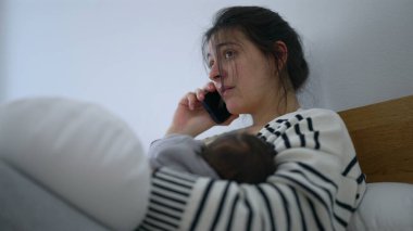 Anne, yeni bebeği kollarında uyurken telefonda konuşuyor. Yorgunluk ve bağlılık karışımını iletiyor. Erken ebeveynlikte dinlenme ve sorumluluğun dengelendiği sakin anlar.