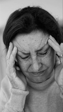 Yaşlı kadın gözlerini kapatıp şakaklarına masaj yapıyor, şiddetli baş ağrısı belirtileri gösteriyor. Onun acı ifadesine odaklan.