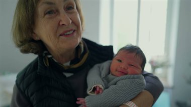 Büyükanne yeni doğmuş bir bebeği kucağında tutuyor, nazik bir gülümsemeyle, sevgi ve bağlılık anını yakalıyor, nesiller boyunca aile bağlarını ön plana çıkarıyor.