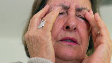 Kıdemli kadın gözlerini sıkıca kapatıyor ve iki eliyle tapınaklara masaj yapıyor. Acıyı ya da stresi azaltmaya odaklanıyor. Kırışıklıklar ve ifade, duygusal yükü vurgular