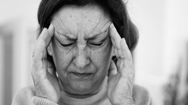 Görünür bir sıkıntı içindeki yaşlı kadın, iki eliyle yüzünü buruştururken, gözleri sımsıkı kapalı, yoğun bir şekilde ağrı kesiciye odaklanmış. Kırışıklıklar ve yüz ifadesi, baş ağrısı veya stres