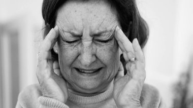 Görünür bir sıkıntı içindeki yaşlı kadın, iki eliyle yüzünü buruştururken, gözleri sımsıkı kapalı, yoğun bir şekilde ağrı kesiciye odaklanmış. Kırışıklıklar ve yüz ifadesi, baş ağrısı veya stres SD 480p
