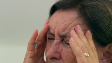 Kıdemli kadın gözlerini sıkıca kapatıyor ve iki eliyle tapınaklara masaj yapıyor. Acıyı ya da stresi azaltmaya odaklanıyor. Kırışıklıklar ve ifade, duygusal yükü vurgular