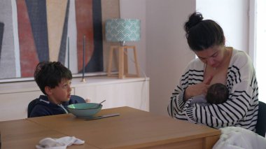 Anne yeni doğan bebeği emzirirken, büyük oğlu yemek masasında oturuyor, elleri kasesinin üzerinde, düşünceler içinde kaybolmuş, sessiz, düşünceli bir aile anını sergiliyor.