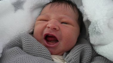 Yeni doğmuş bebek sıcak bir battaniyeye sarılmış, hafif kaşları çatık, rahatsızlık belirtileri gösteriyor, erken dönem duygularını yakalıyor, sıcak ve güvenli bir şekilde sarmalanıyor.