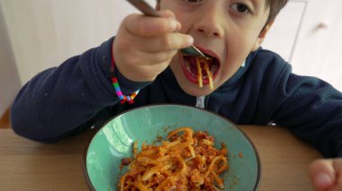 Çocuk yemek yerken spagettiyi dikkatlice çeviriyor, kararlılığını gösteriyor ve yemeğe odaklanıyor, aşina olduğu bir mutfak ortamında kendini besleme sürecini vurguluyor.