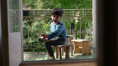 Genç çocuk bir balkonda tahta bir taburede oturuyor, gülümsüyor ve ayağını tutuyor, yeşilliklerle çevrili, yumuşak doğal ışıkla.