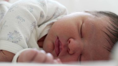 Serene uyuyan bebek, beyaz tulum içinde, bebeğin yüzünün ve elinin yakın çekimi, erken çocukluğun yumuşaklığını ve saflığını yakalıyor.