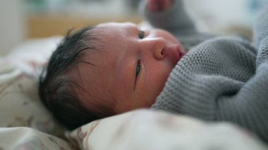 Yeni doğmuş bebek, geniş gözlerle uzanmış, rahat bir battaniyeye sarılmış, meraklı bir şekilde dünyaya bakıyor, masumiyetini ve erken yaşamın mucizesini vurguluyor.