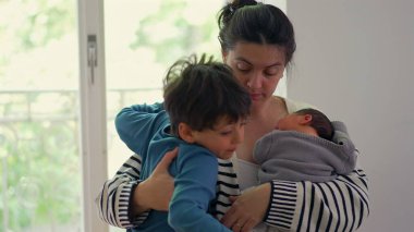 Anne hem yeni doğan hem de büyük çocuğu kucaklıyor, hassas bir aile anında kardeşler arasındaki sevgi ve ilgiyi dengeliyor, anne olmanın önemini vurguluyor.