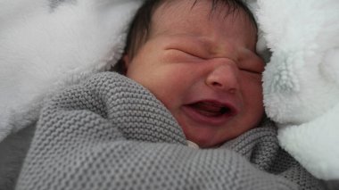 Yeni doğmuş bebek sıcak bir battaniyeye sarılmış, hafif kaşları çatık, rahatsızlık belirtileri gösteriyor, erken dönem duygularını yakalıyor, sıcak ve güvenli bir şekilde sarmalanıyor.